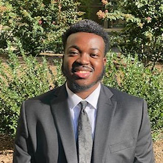 A young man in a suit smiles against a background of trees and green plants.