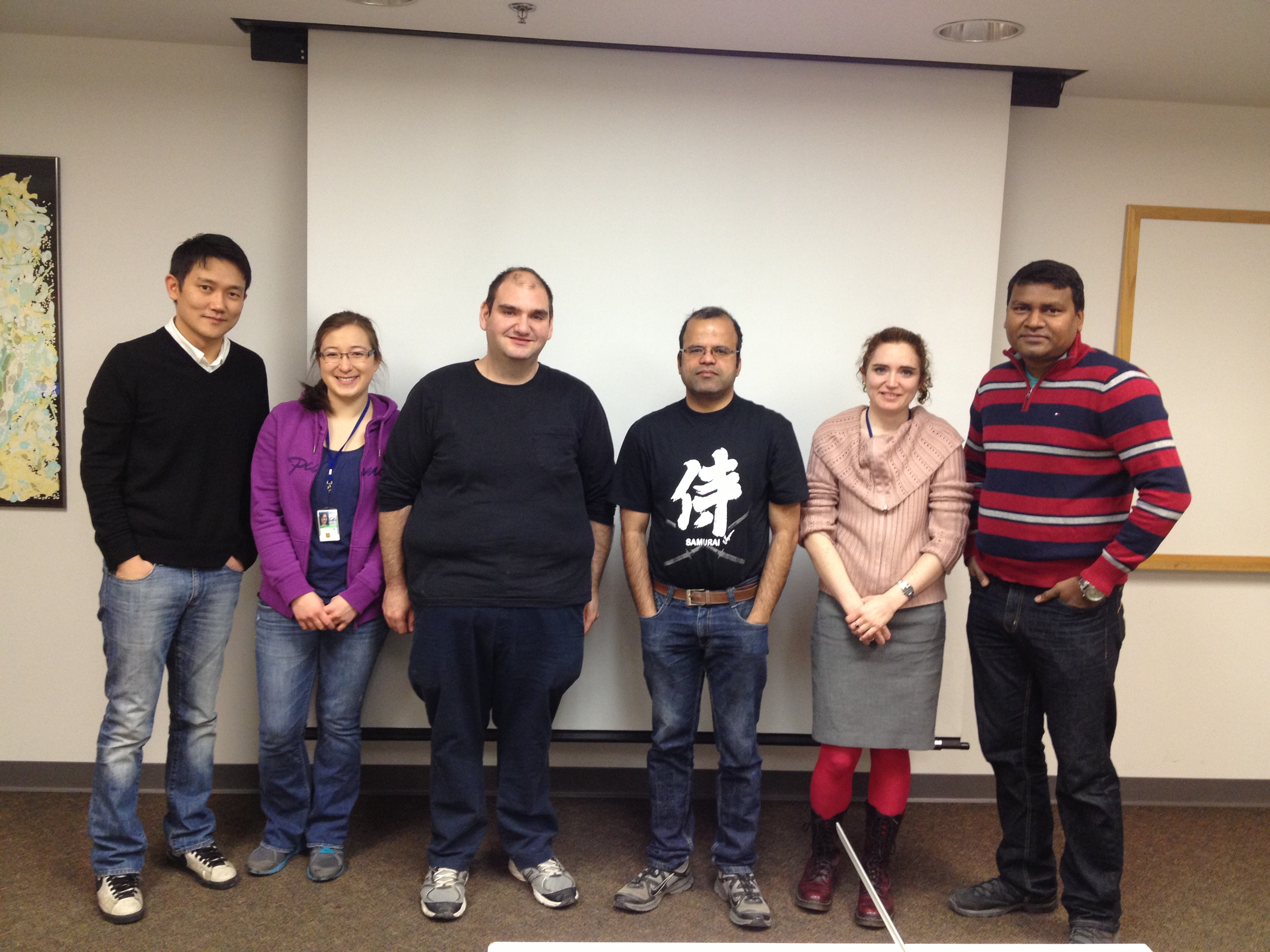 Six lab members pose in a line in front of a projection screen in building 32TII.