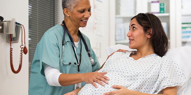 A pregnant woman sits up on a hospital bed and is speaking with a health care provider.