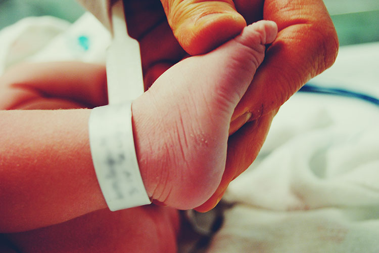 A newborn’s foot, with a hospital ankle tag, is held by an adult hand.