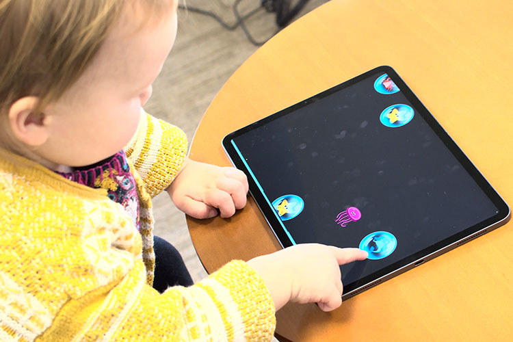 A toddler wearing a yellow sweater and seated at a table uses her fingertip to pop a bubble on a tablet device.