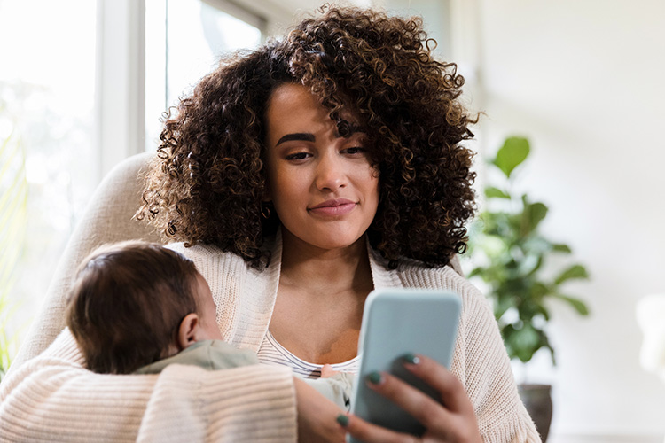 A mother holds her baby while looking at a cell phone.