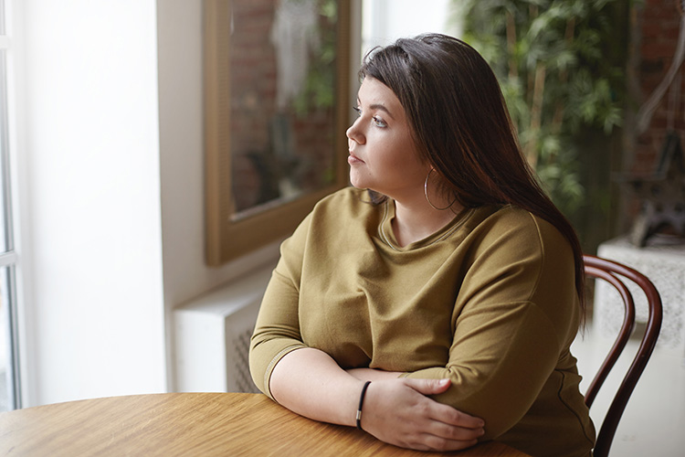 A young woman with overweight sitting at a table and looking through a window. 
