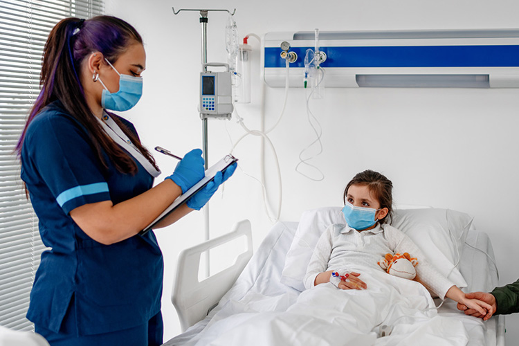Child lying in hospital bed holding caretaker’s hand. Health care worker writing on clipboard. Both are masked. 
