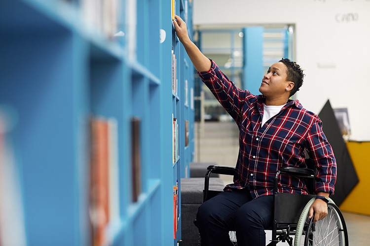 A person in a wheelchair reaches for a book on a library shelf.