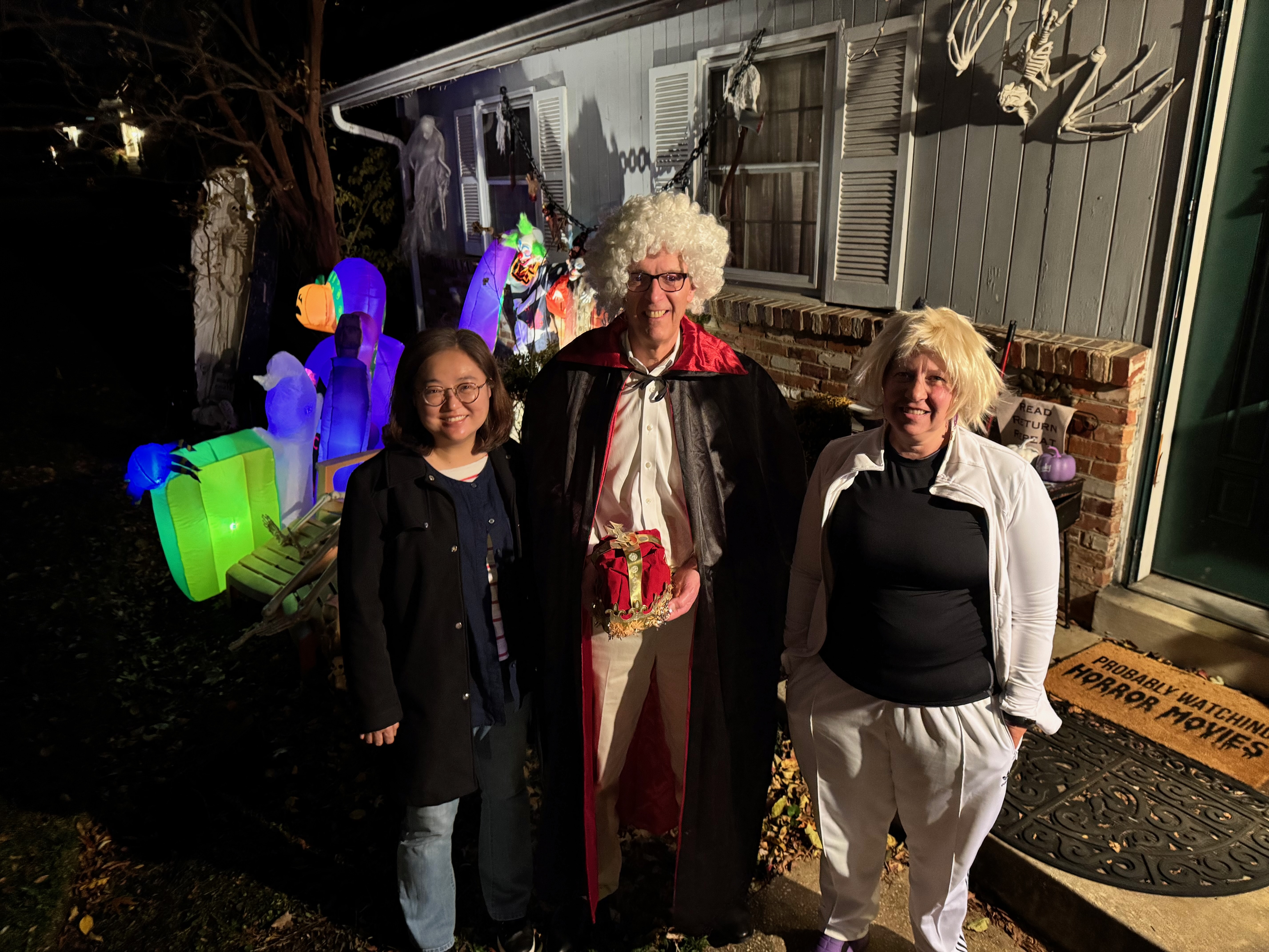 Three lab members in costumes pose together outside a decorated house, celebrating Halloween 2025. 