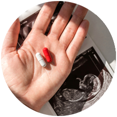 A red pill and a white pill rest in the palm of a hand extended above a fetal ultrasound printout. 