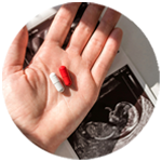 A red pill and a white pill rest in the palm of a hand extended above a fetal ultrasound printout. 