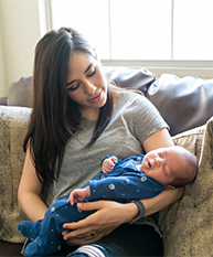 A young woman sits on a sofa and looks at her baby who is asleep in her arms.