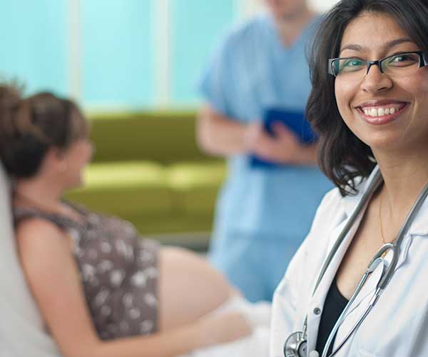 Female doctor in foreground smiling; in the background, a pregnant woman sitting on an exam table speaks to a medical professional.