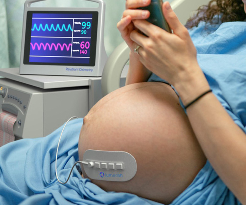 A pregnant women in a healthcare setting has the Lumerah oximeter on her belly while she uses a smartphone.