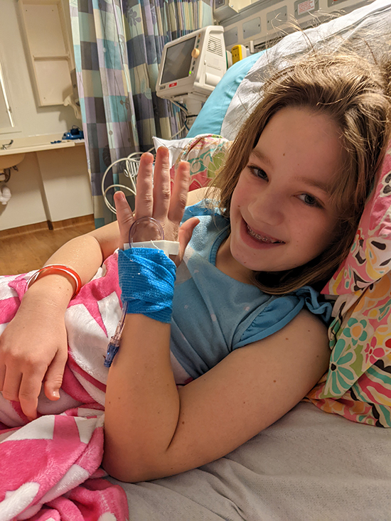 A smiling young girl wearing a blue shirt lies in a hospital bed. She holds up her hand, which is covered by a blue bandage holding an IV in place.