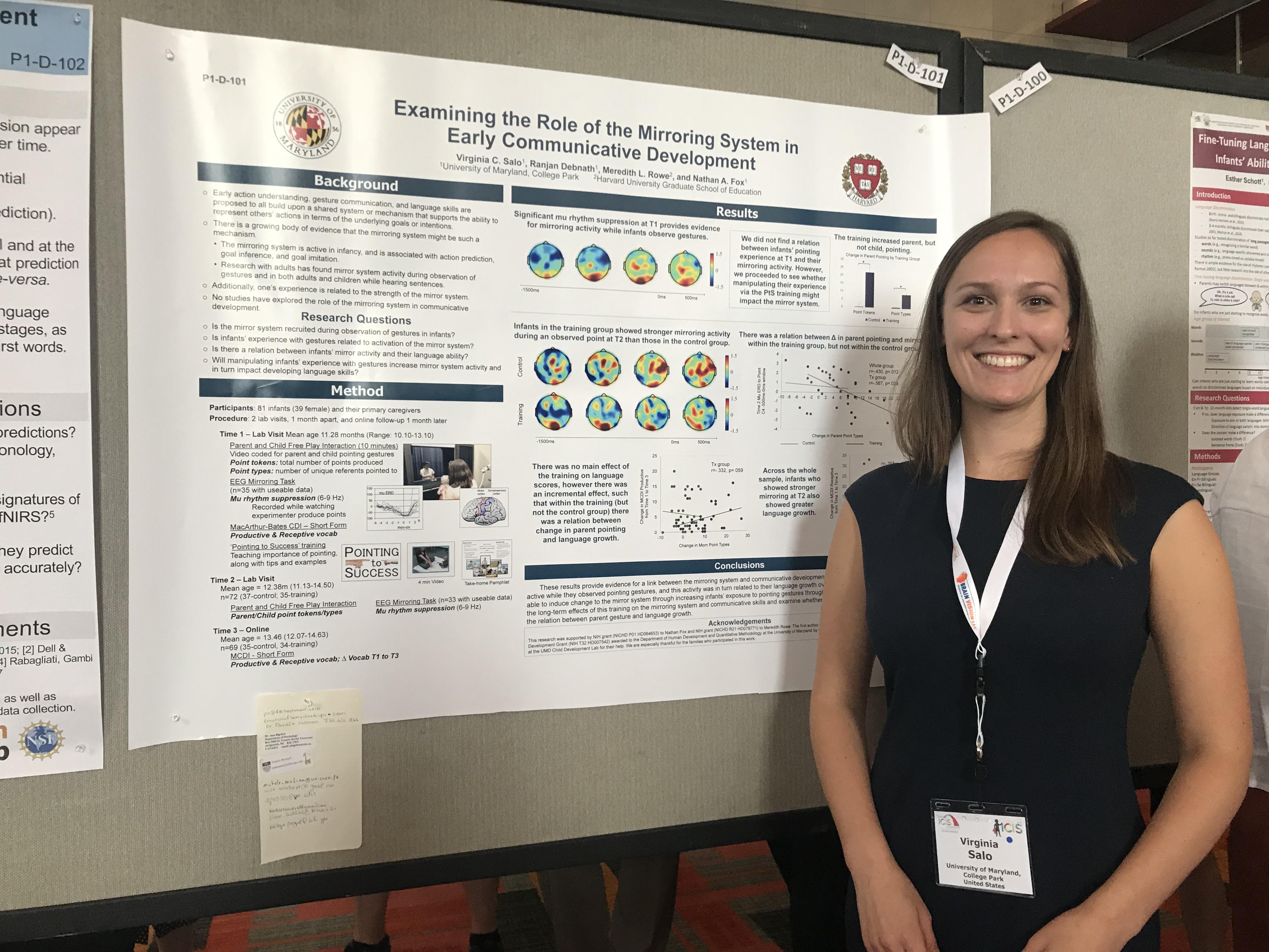 A smiling woman wearing a black shirt and white name badge stands beside a scientific poster titled 'Examining the Role of the Mirroring System in Early Communicative Development.'