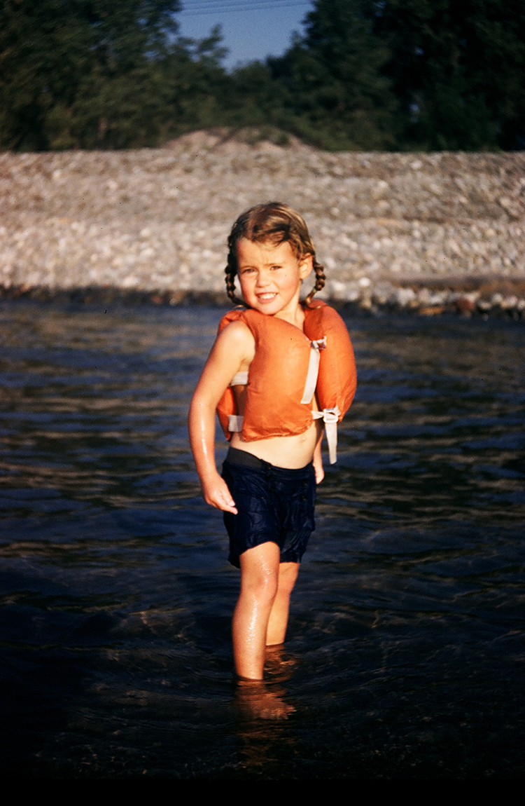 A young girl wearing a life vest stands smiling in ankle-deep water.
