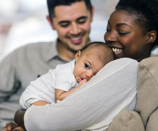 A man and woman sit together on a couch as the woman cradles a baby in her arms. Both are smiling at the baby.