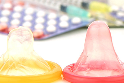 Three condoms, with birth control pills and other forms of contraception in the background.