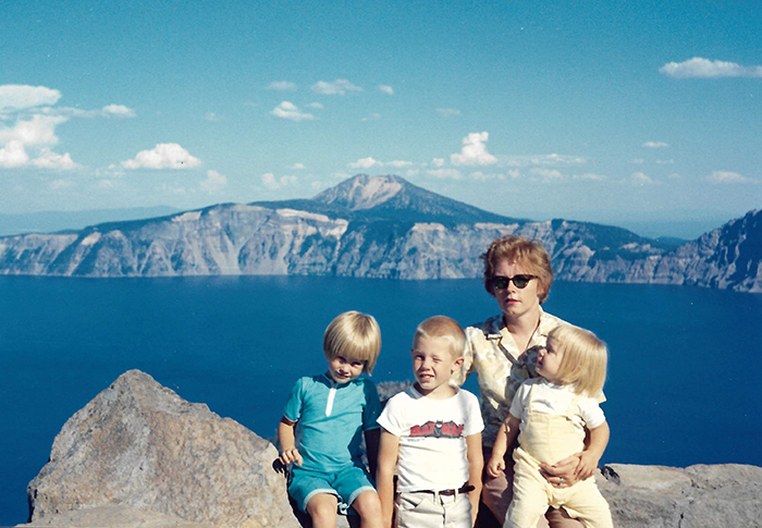 Three blond-haired children are sitting with their mother, who is wearing sunglasses, in front of Crater Lake.