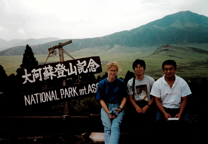 The  group is sitting next to a wooden sign that has white letters. The top of the  sign has Japanese writing and the bottom says, &ldquo;NATIONAL PARK mt ASO.&rdquo; The  mountain is visible in the background. 