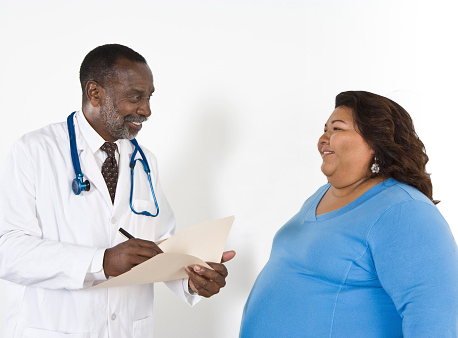 Un proveedor de servicios de salud con un estetoscopio y escribiendo en una tabla al mismo tiempo que habla con una mujer embarazada.