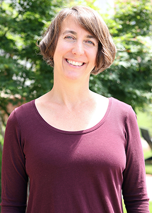 A woman smiles in an outdoor setting with trees and grass.