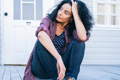 A woman sits on the steps of a deck outside a house with her knees drawn up close to her body. She rests her head in the palm of one hand as she stares into the distance.
