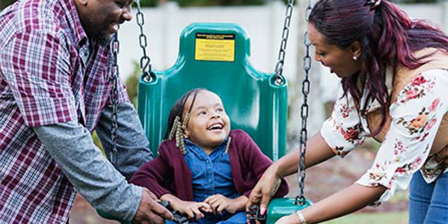 An African-American family at the playground with a happy 5 year old daughter. The little girl has caudal regression syndrome, a rare congenital disorder which affects the development of the lower spine. She is smiling as her parents buckle her into a swing.