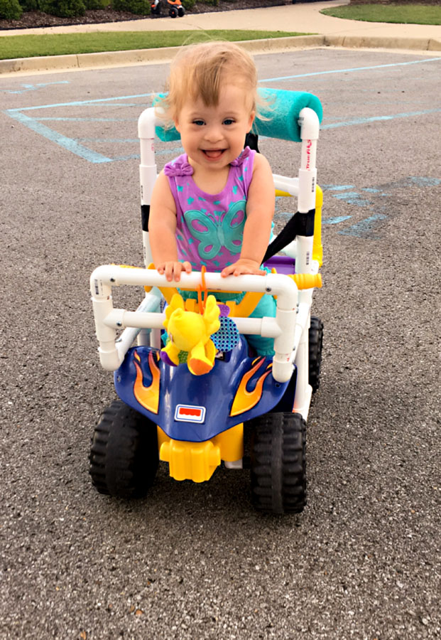A toddler girl with Down syndrome riding in a child’s toy car.
