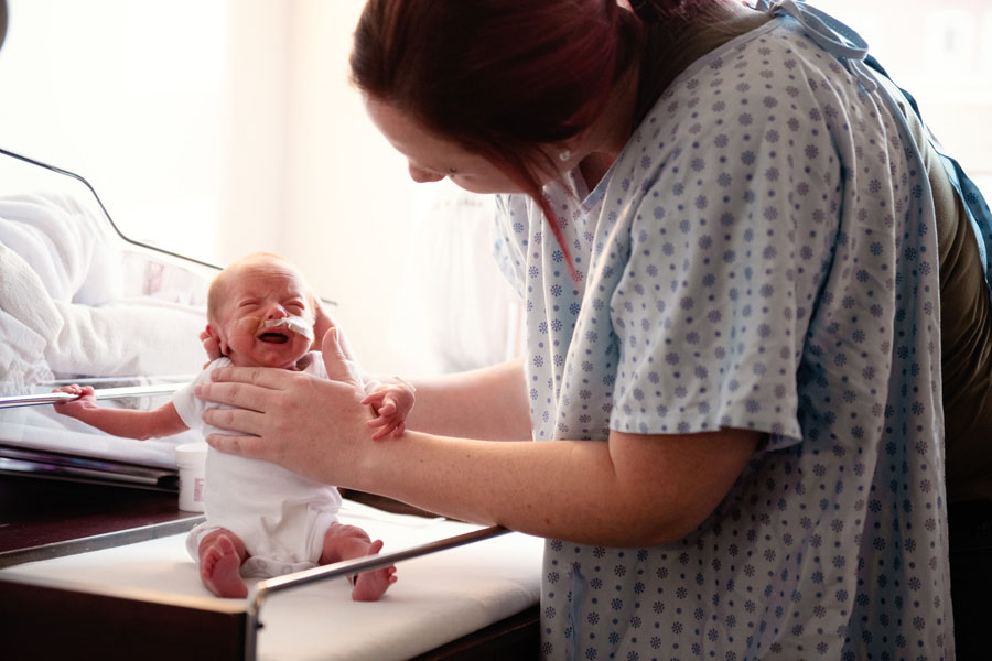 A woman in a hospital gown supporting newborn seated on a portable hospital crib. The infant has a breathing tube attached to its nose.