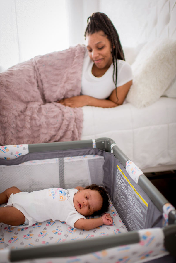 A mother lying in bed looking down at a baby lying on its back in a crib next to the bed.