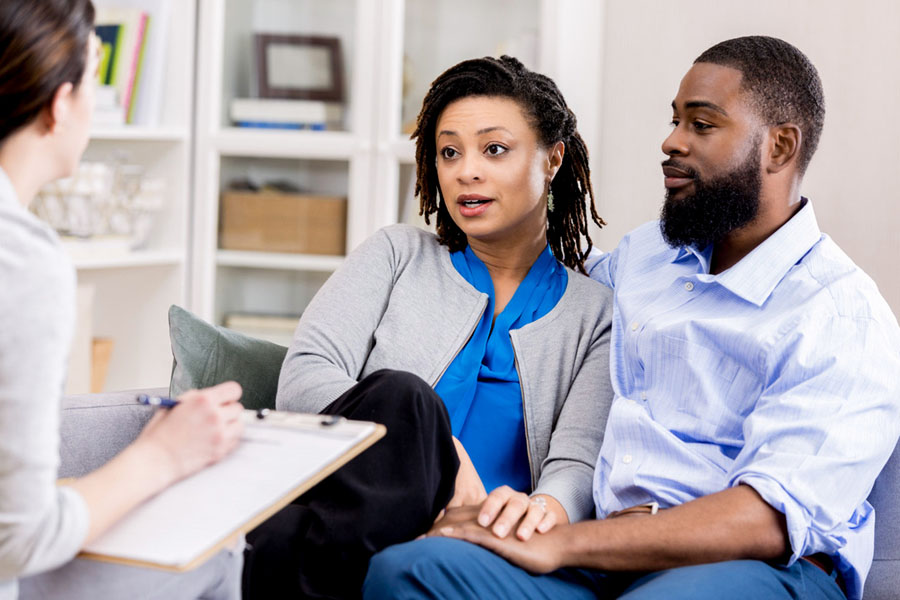 A man and woman seated together and holding hands as they talk with a professional holding a clipboard.