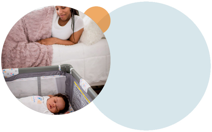 A mother lying in bed looking down at a baby lying on its back in a crib next to the bed.