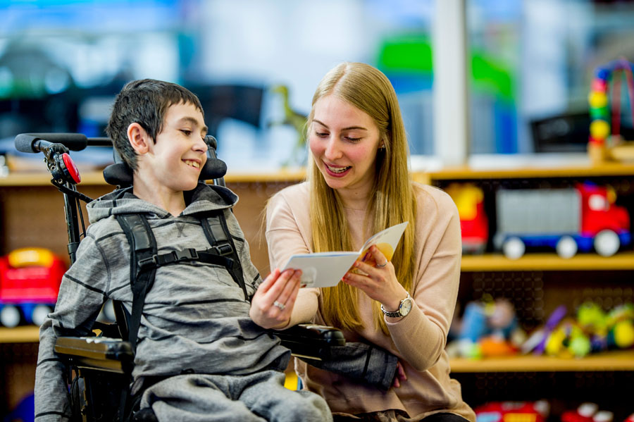 A young woman reading a book to a child in a wheelchair. 