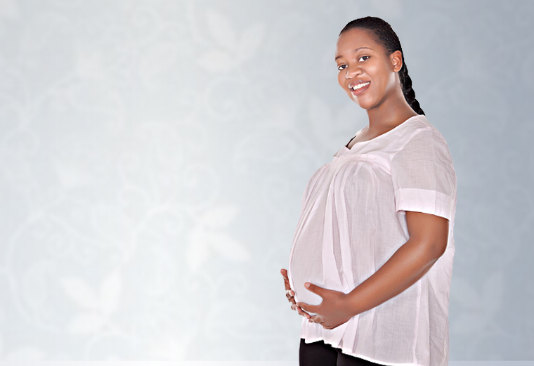 A smiling pregnant woman in a maternity blouse holding her abdomen.