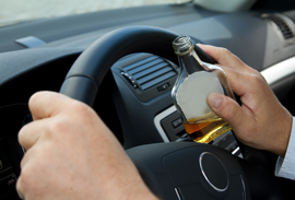 Hands holding car steering wheel while clutching liquor bottle.