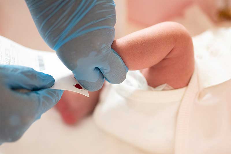 Gloved hands holding infant heel, blood spot visible on filter paper.